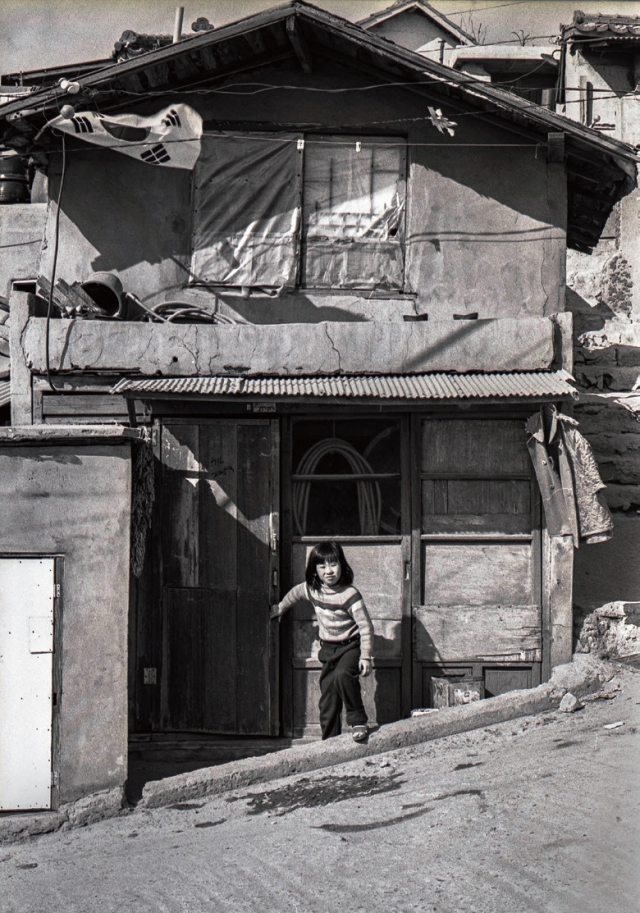 김정일, 기억 풍경-봉천동(1982). 경사로에서 곧장 집으로 이어지는 산동네 특유의 건축 구조를 가진 집에서 한 아이가 나오고 있다.