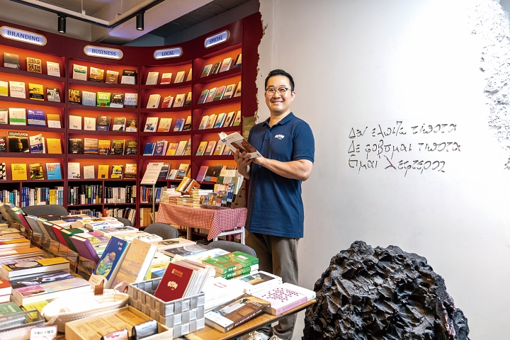 책방 &lsquo;크레타&rsquo; 강동훈 대표가 자신의 인생책 &lsquo;그리스인 조르바&rsquo;를 손에 든 채 활짝 웃고 있다. 자줏빛 색감의 책장, 돌 조형물, 벽면에 새긴 니코스 카잔차키스의 묘비명 등은 모두 소설 속에서 영감을 얻었다. 사진 C영상미디어