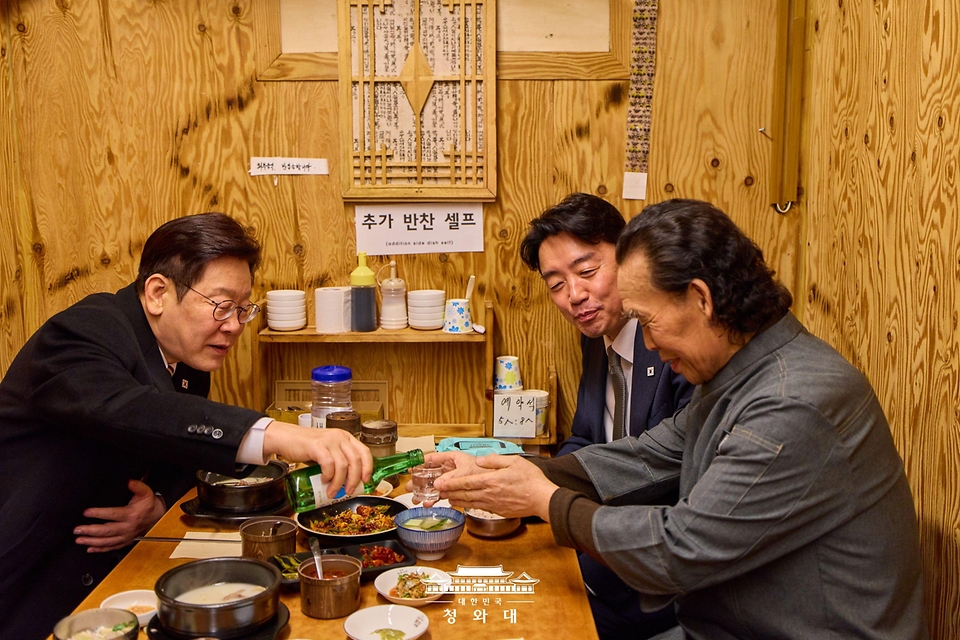 이재명 대통령이 2월 9일 서울 종로구 통인시장 한식당에서 강훈식 대통령비서실장과 저녁 식사를 하며 식당 주인에게 술을 권하고 있다.