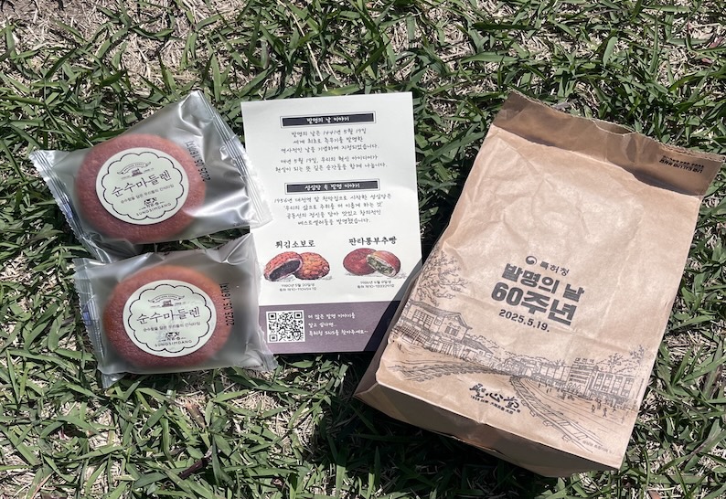 잔디 위에 놓인 성심당 순수 마들렌 2개, 발명의 날·지식재산 리플릿, 기념 종이봉투 구성물.