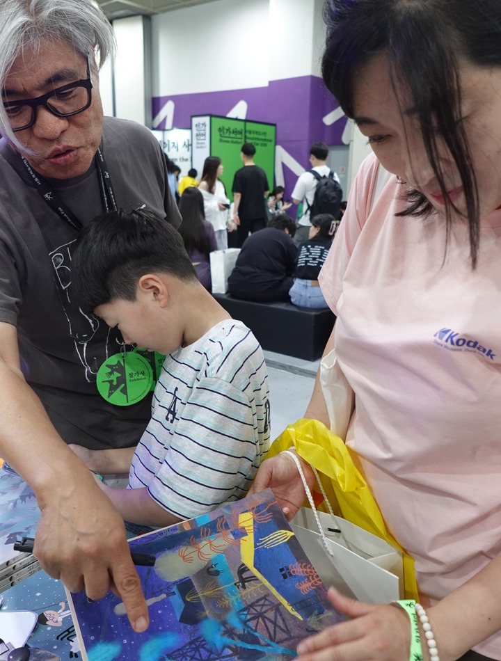 아동 도서 이경국 작가(왼쪽)가 어린이와 보호자에게 그림책을 소개하며 작품을 설명하고 전주에서 온 블로그 운영자이자 작가 최미리 씨와 아들 이상윤 군이 2026 서울국제도서전에서 어린이 도서를 살펴보고 있다. (본인 촬영)