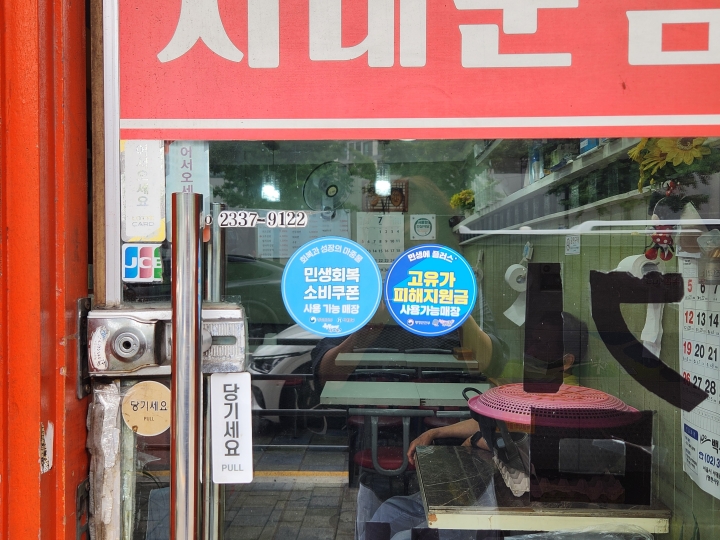 매장 출입문에 지난해 '민생회복소비쿠폰'과 올해 '고유가 피해지원금' 사용 가능 스티커가 함께 붙어 있다. 정부 소비지원 정책이 지난해부터 이어지고 있음을 보여준다. (본인 촬영)