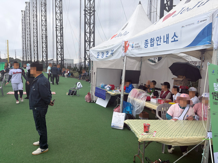 야구 경기장 주변 자원봉사센터에서 분홍색 옷과 모자를 착용한 봉사자들이 자리를 지키고 있다. 앞에는 경기장 관람 인증 행사 안내판이 세워져 있다. (본인 촬영)